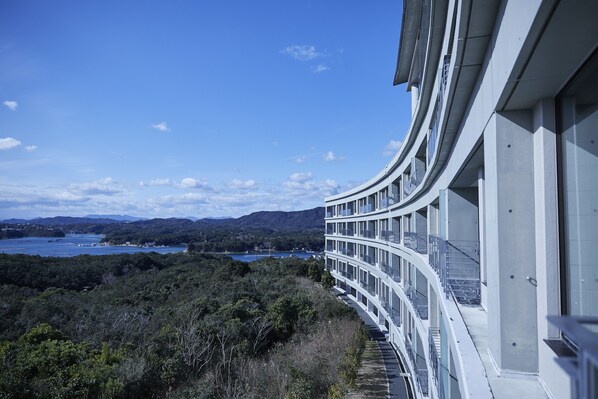 A modern building with a curved balcony overlooking a coastal landscape with mountains and a body of water.