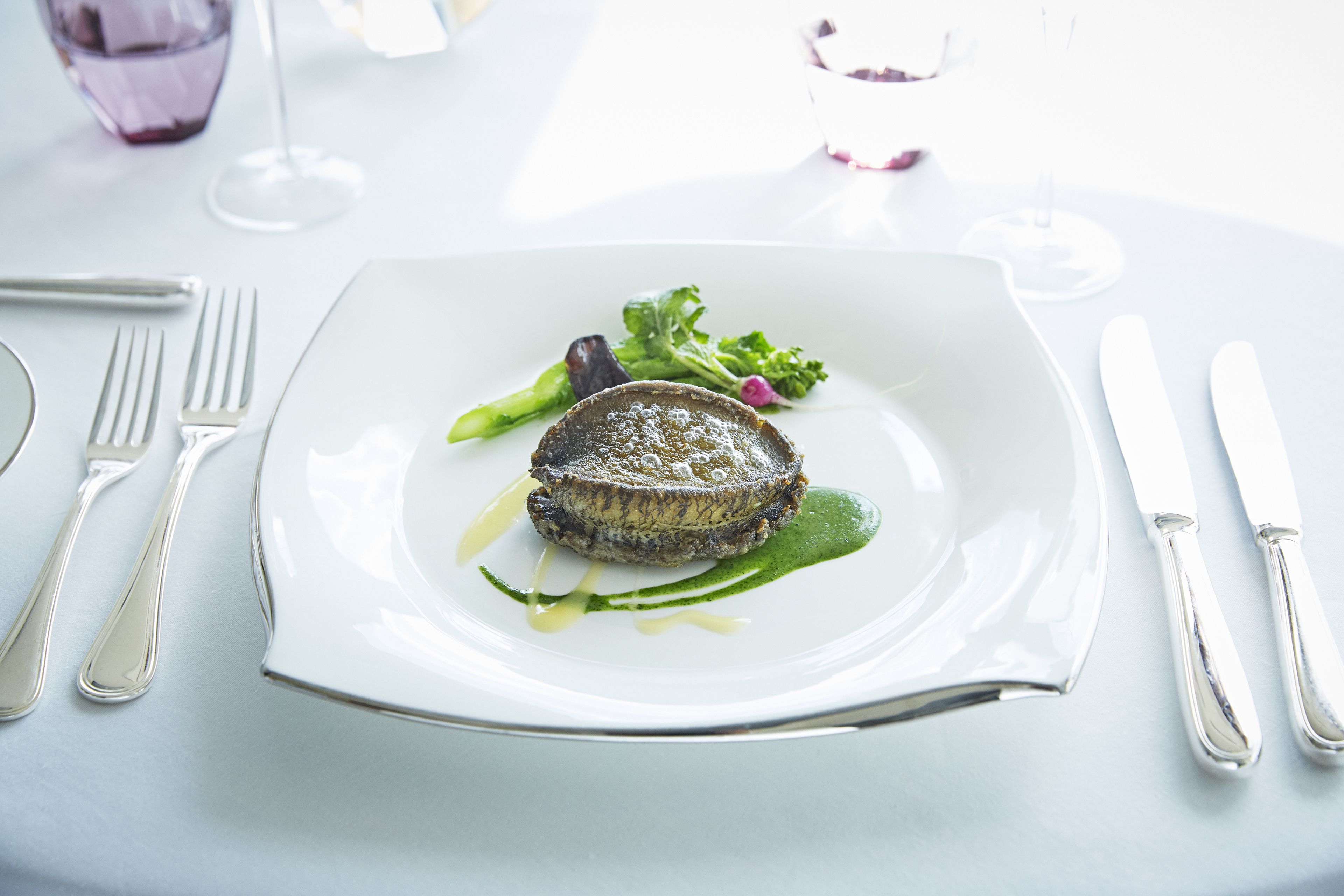 A white plate with a steak, green sauce, and vegetables on a table with a fork, knife, and glass.