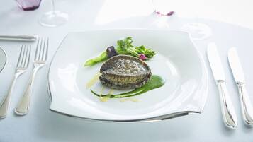 A white plate with a steak, green sauce, and vegetables on a table with a fork, knife, and glass.