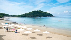 A beach with umbrellas, a sandy shore, and a hill in the background. - Shima Kanko Hotel The Bay Suites (Shima)