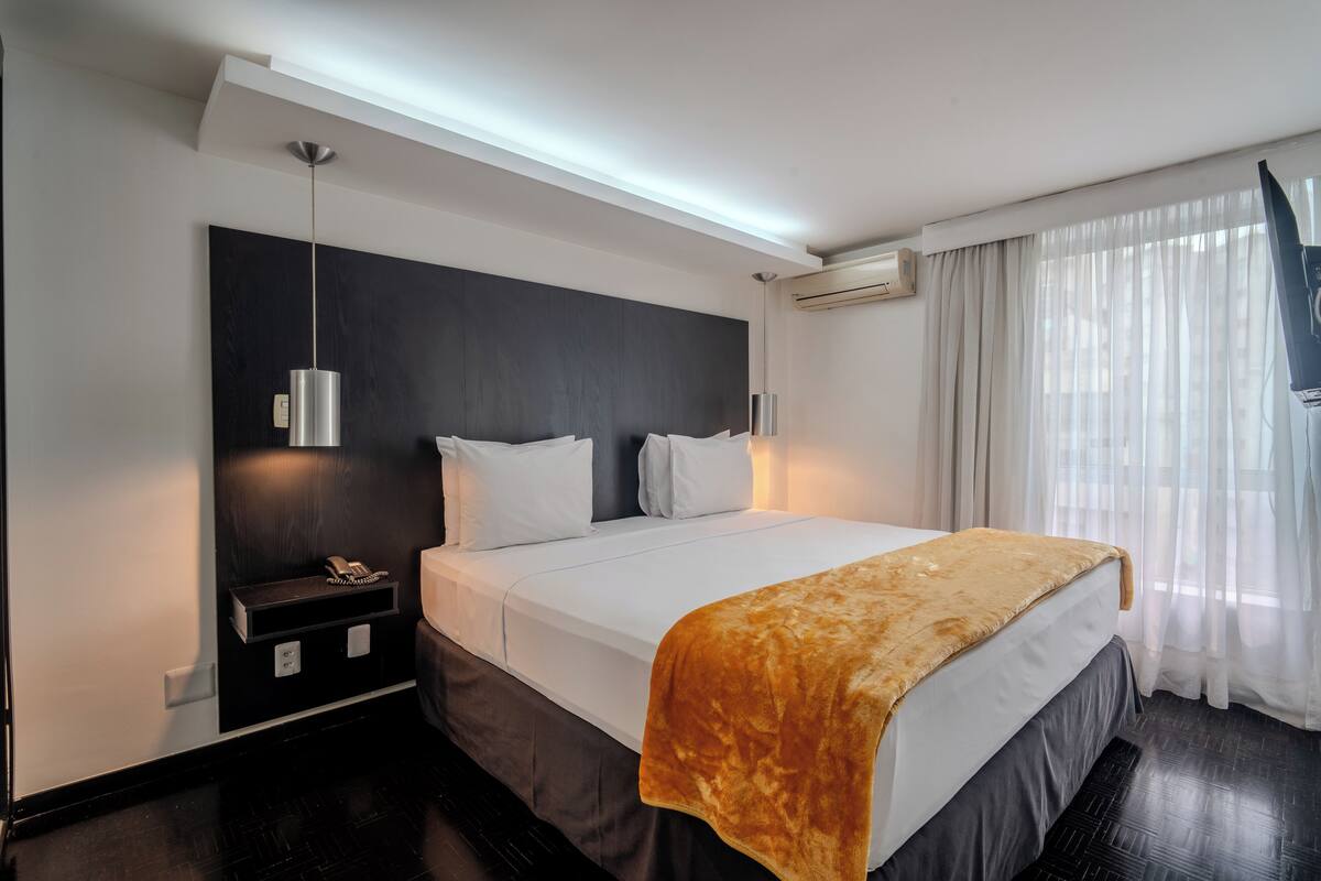 A modern hotel room with a large bed, bedside tables, a hanging light fixture, and a window with sheer curtains.