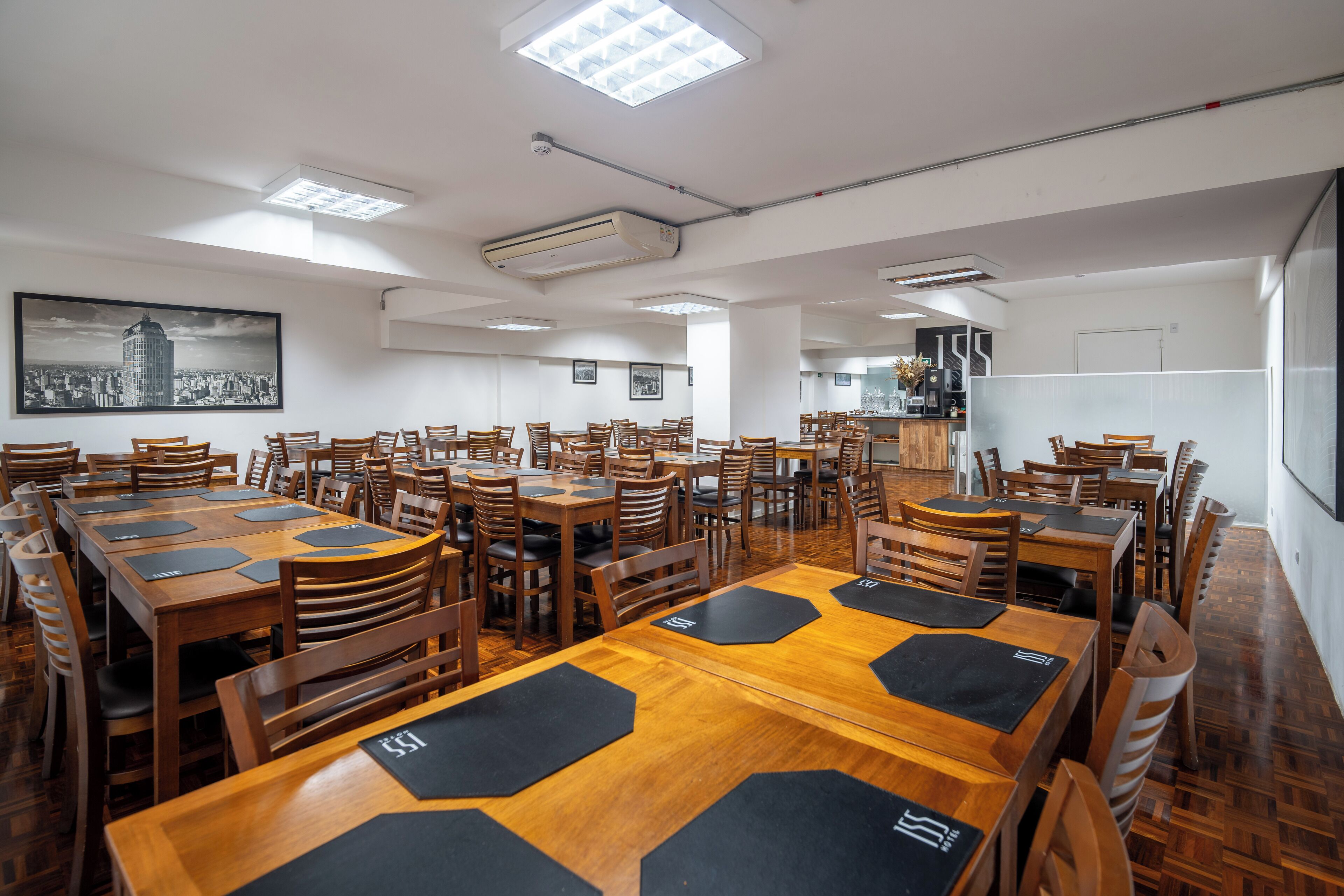 A spacious dining area with multiple wooden tables and chairs, a large cityscape painting on the wall, and a kitchen area in the background.