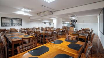 A spacious dining area with multiple wooden tables and chairs, a large cityscape painting on the wall, and a kitchen area in the background.