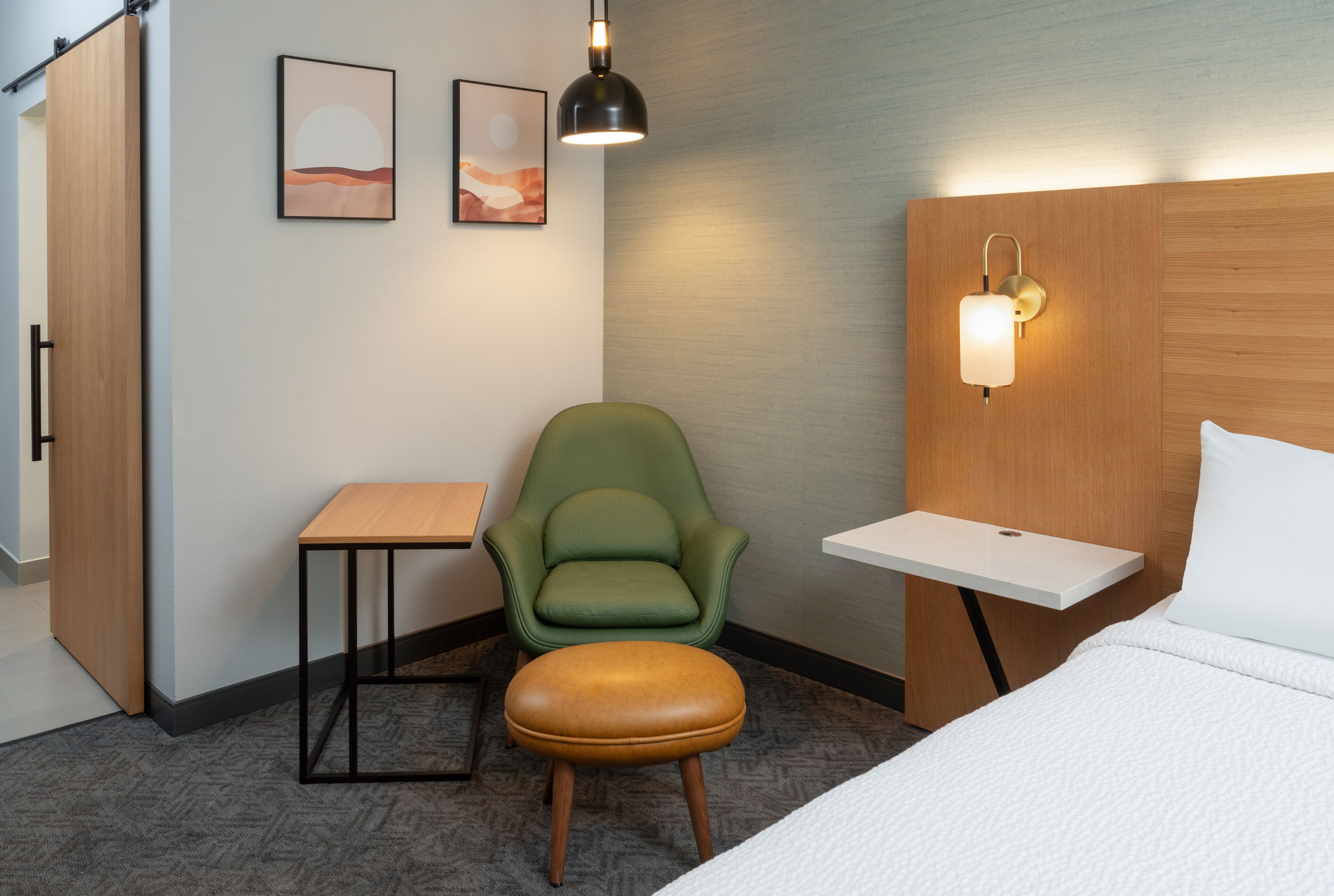 A hotel room with a bed, a green armchair, a small table, a wooden nightstand, and two wall-mounted light fixtures.