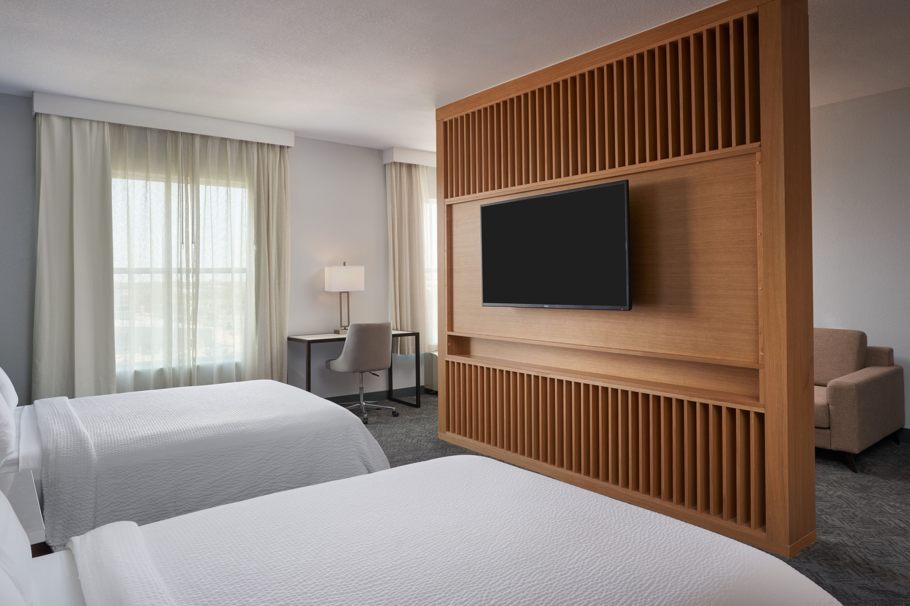 Quarto de hotel com duas camas, uma televisão fixada em um painel de madeira, uma escrivaninha com cadeira e uma janela com cortinas.