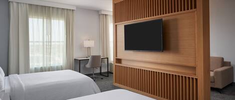 Quarto de hotel com duas camas, uma televisão fixada em um painel de madeira, uma escrivaninha com cadeira e uma janela com cortinas.
