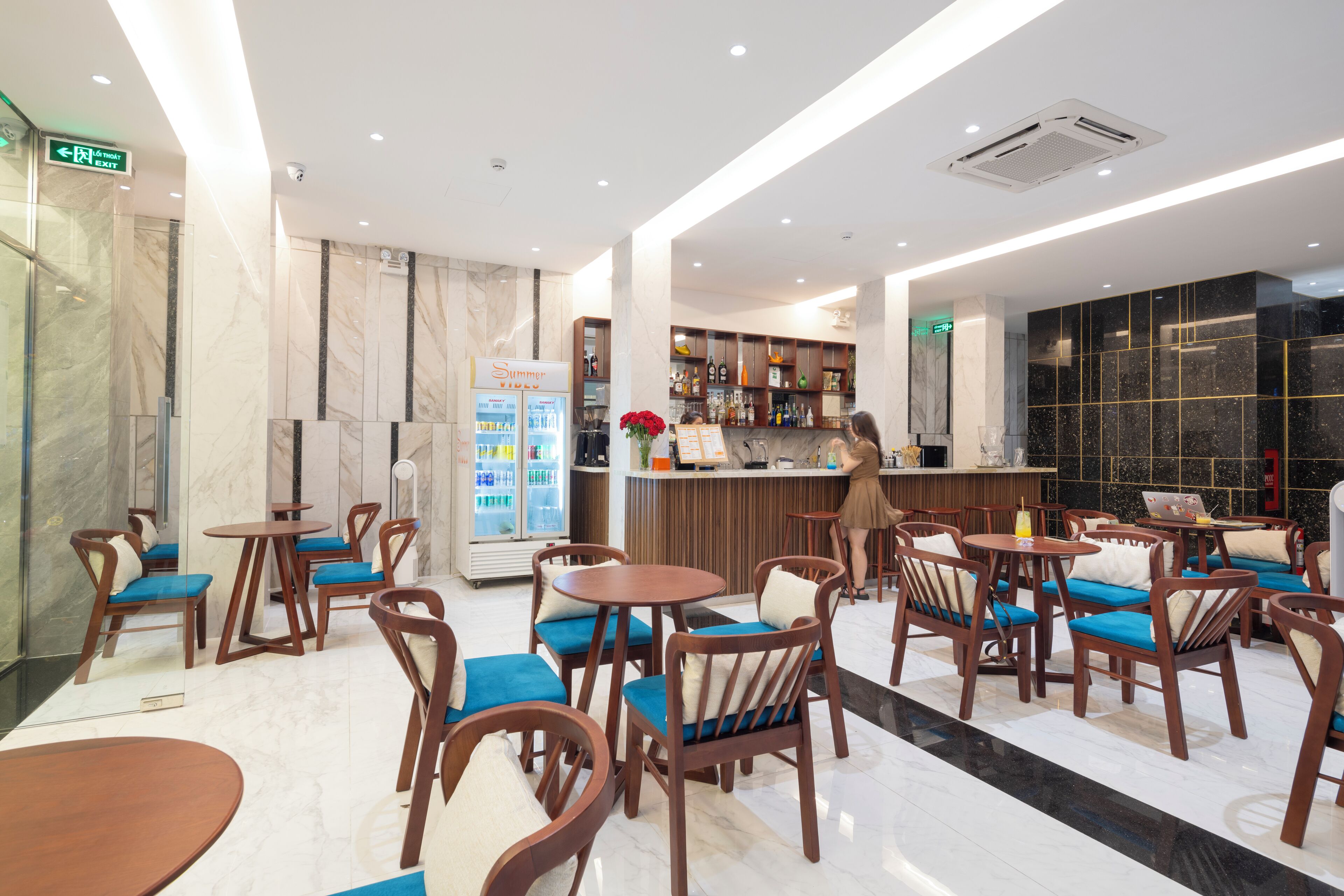A modern hotel lobby with marble flooring, wooden furniture, a bar area, and a refrigerated display.