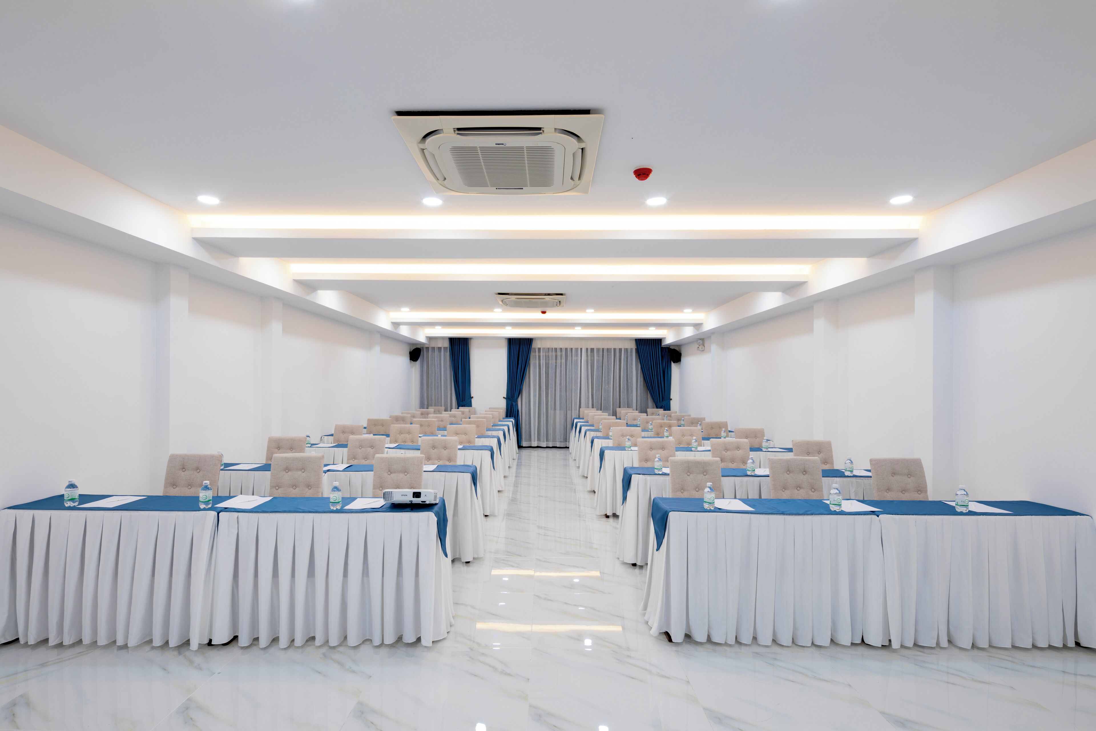 A conference room with rows of tables and chairs, a marble floor, and a ceiling with recessed lighting and an air conditioning unit.