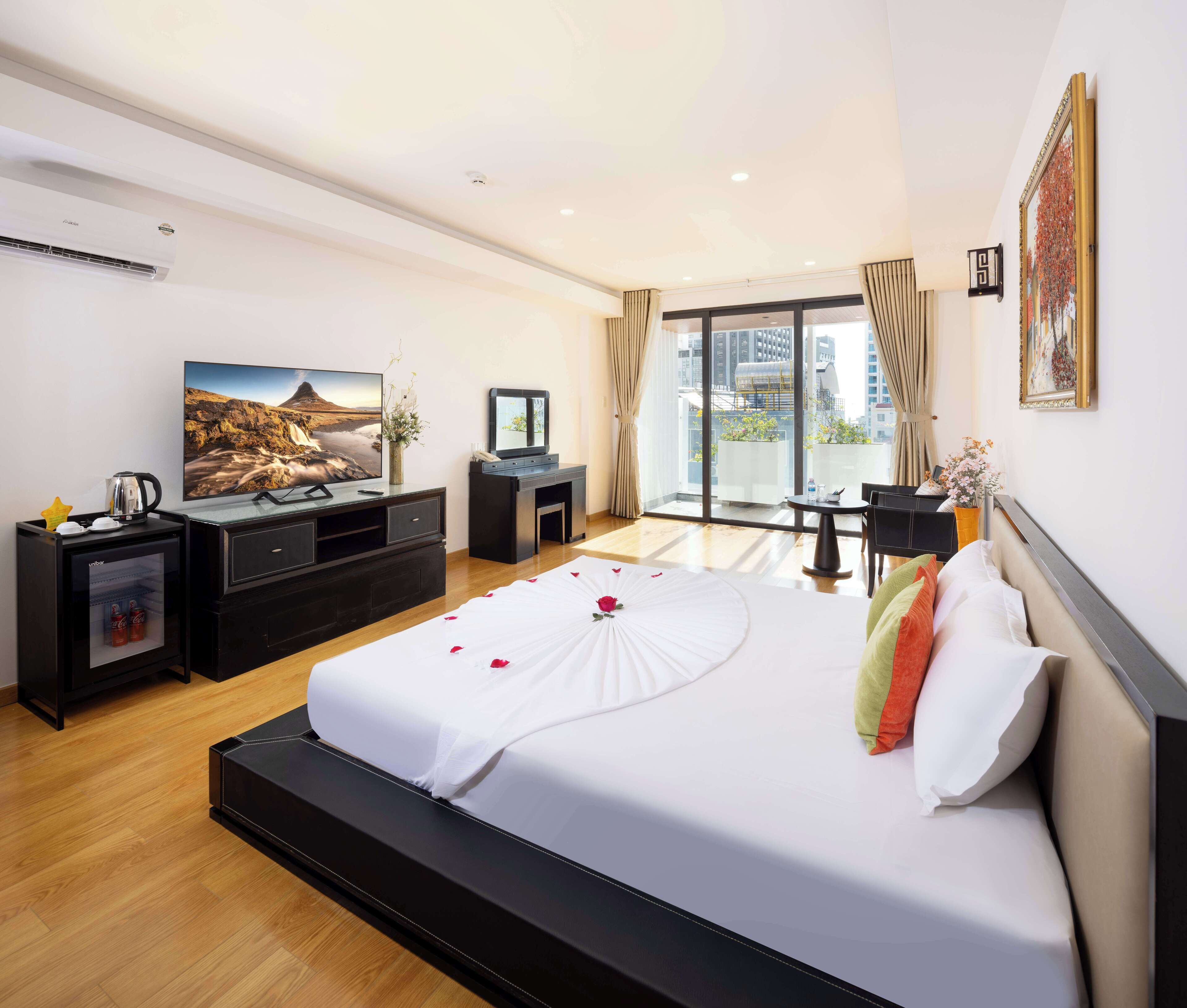A modern hotel room with a large bed, a flat-screen TV, a desk with a chair, a small table, and a balcony with a view of the city.