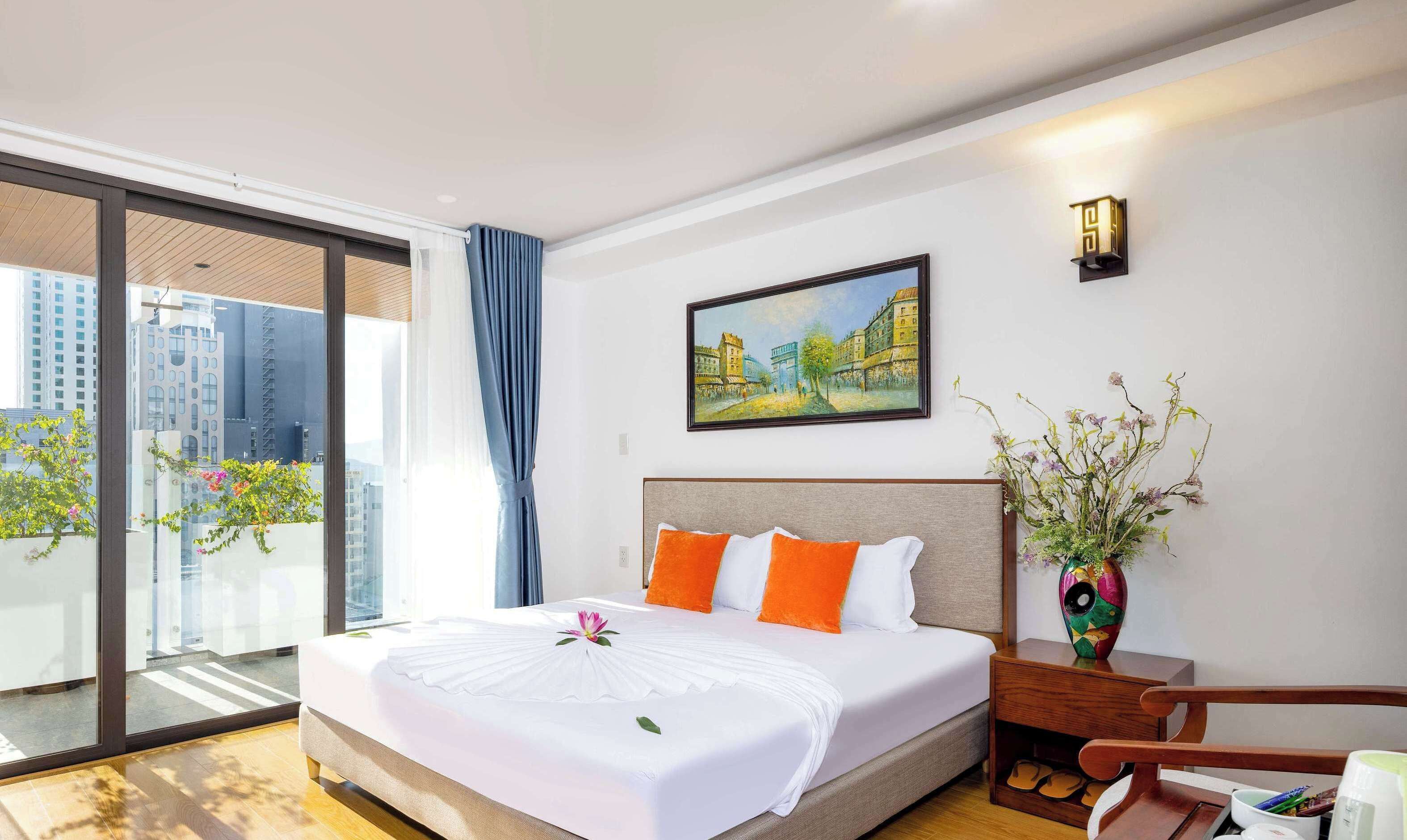 A hotel room with a large bed, a painting on the wall, a balcony with plants, and a view of buildings.