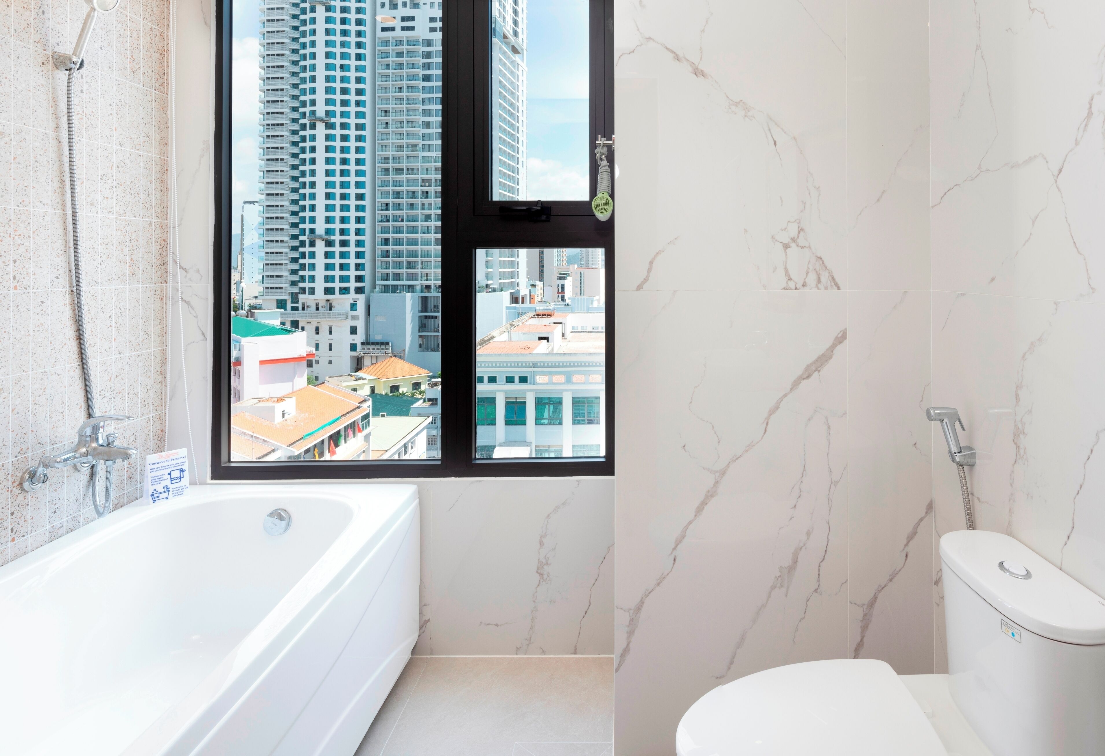 A modern bathroom with a bathtub, toilet, and a window offering a city view.
