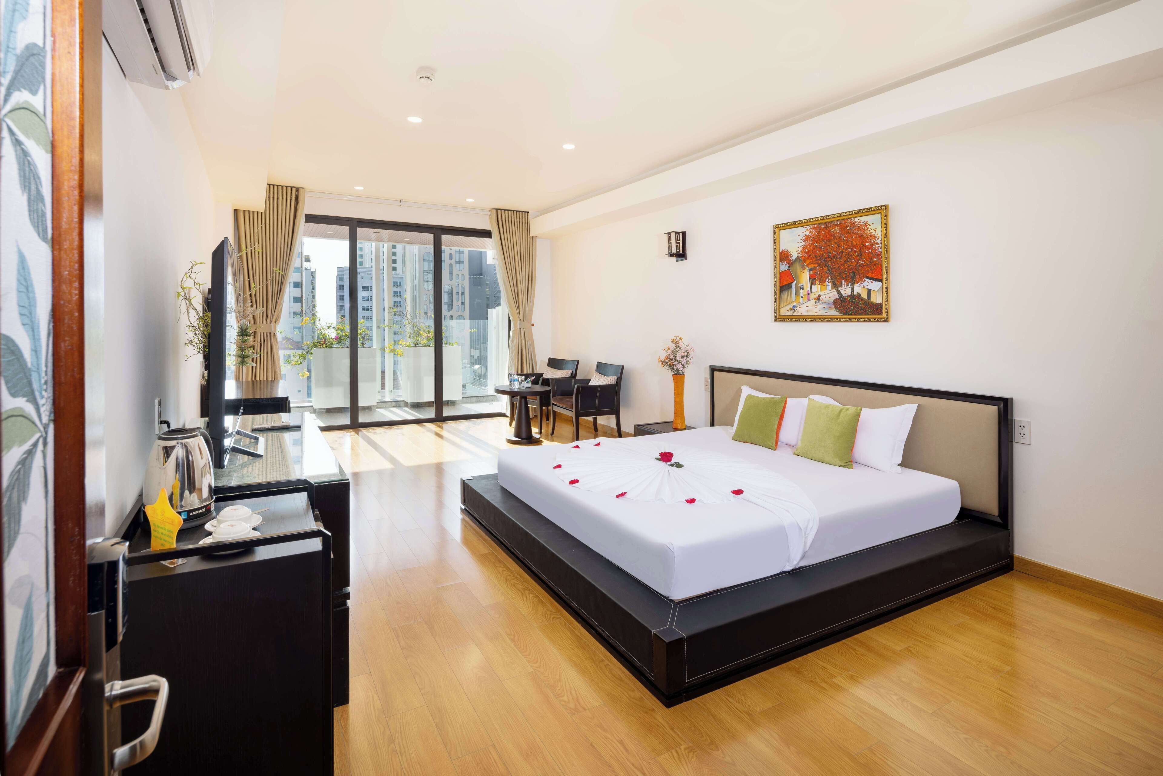 A hotel room with a bed, a desk, a chair, a small table, and a large window with a view of buildings.