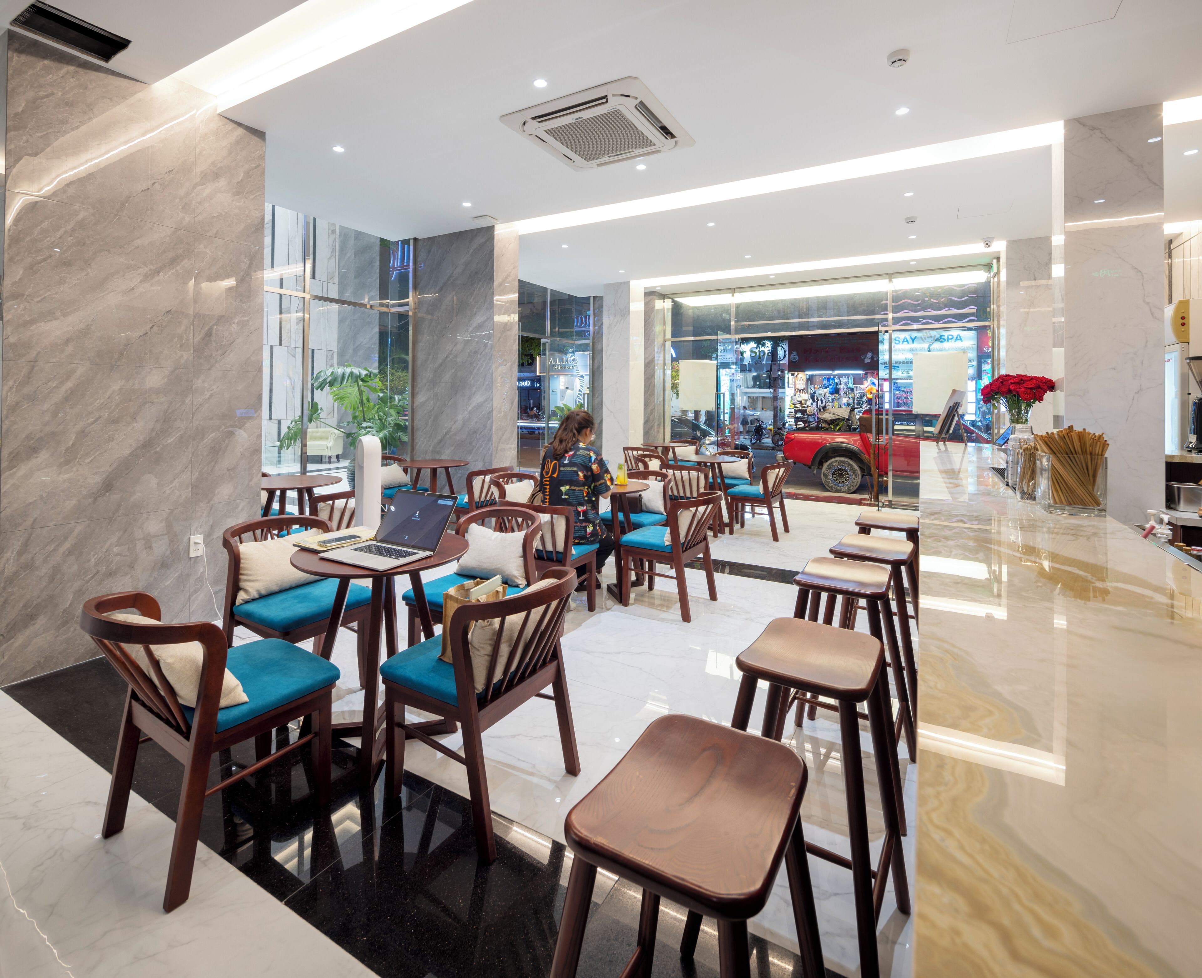 Un café moderno con mobiliario de madera y cojines azules, una pared de vidrio que ofrece vista a un coche rojo en el exterior.