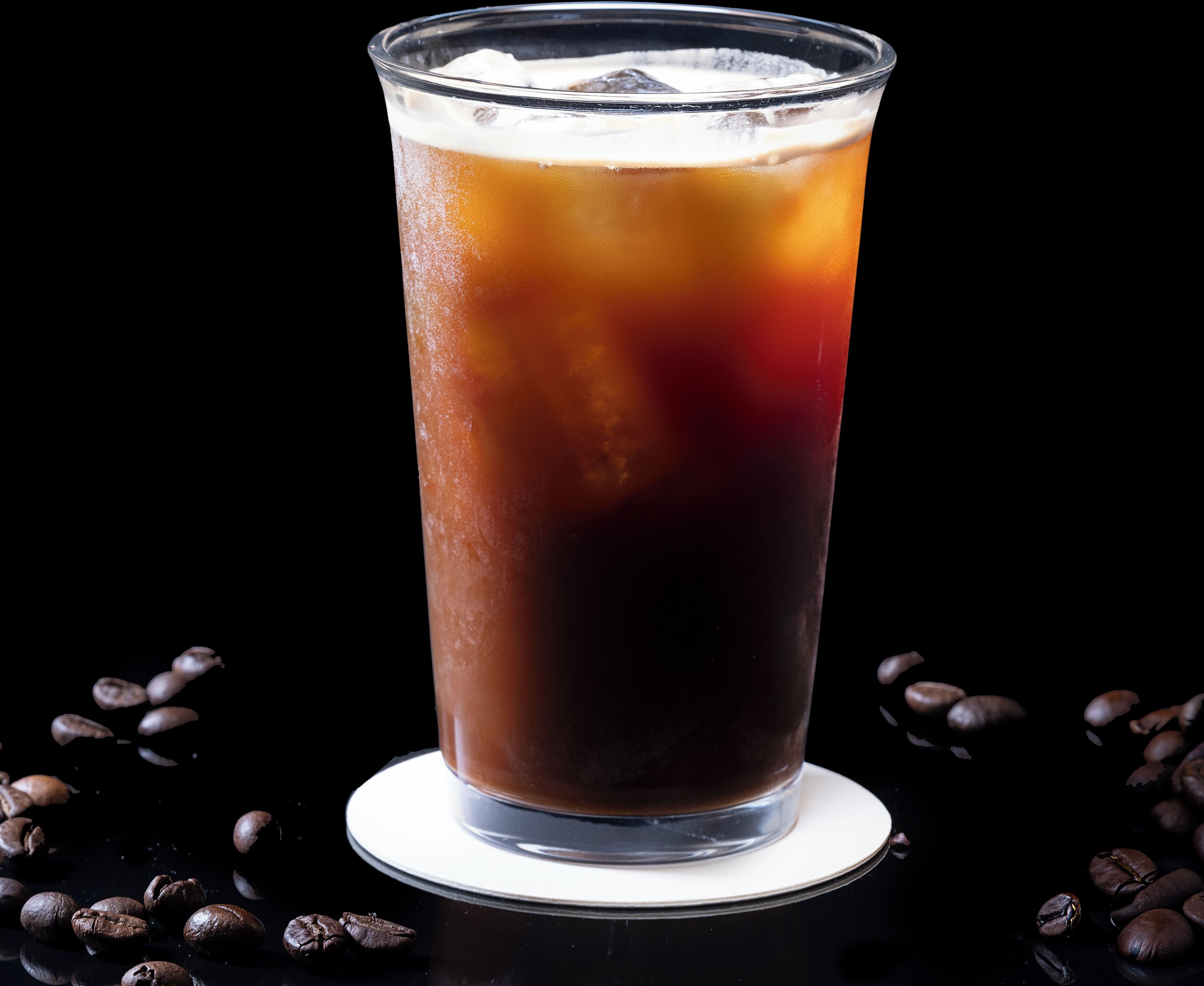 A tall glass of iced coffee with ice cubes, surrounded by coffee beans on a reflective surface.