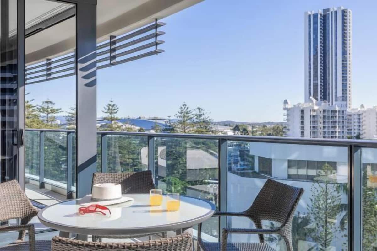 Un balcón con mesa, sillas y barandilla de vidrio con vista a un paisaje urbano.