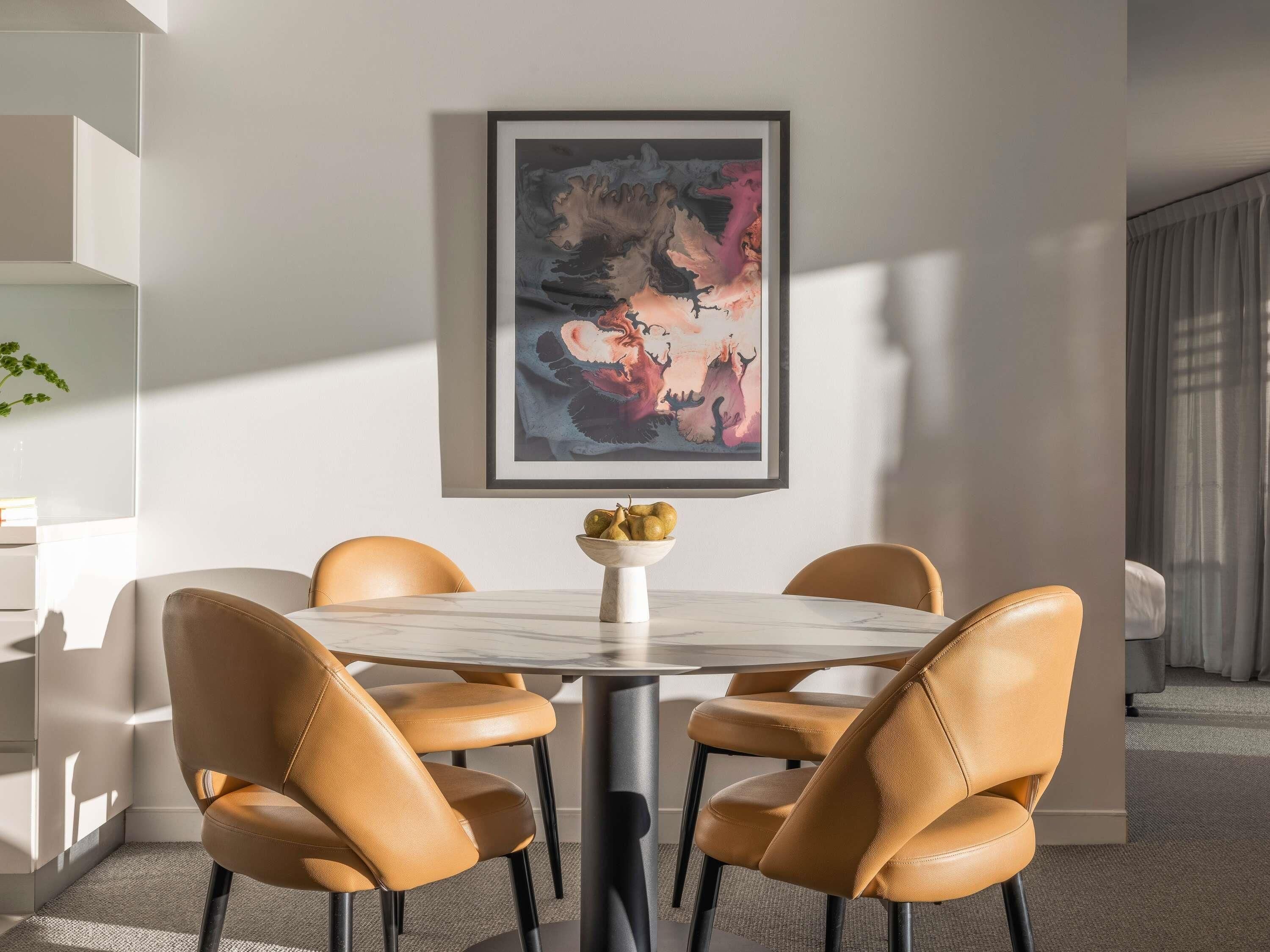 A dining area with a round table, four chairs, a framed abstract painting, and a plant on the left.