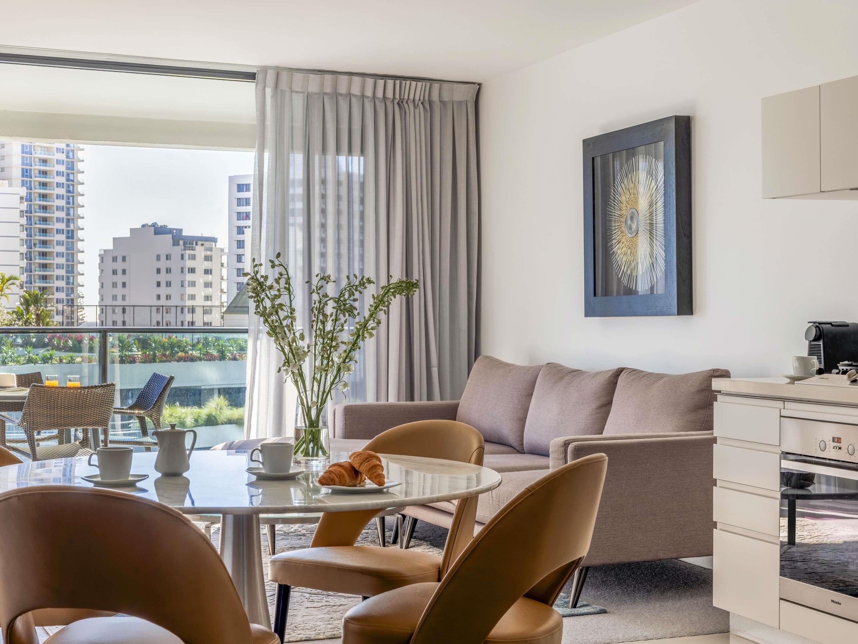 A modern living room with a sofa, chairs, a dining table, a coffee machine, and a view of the cityscape.