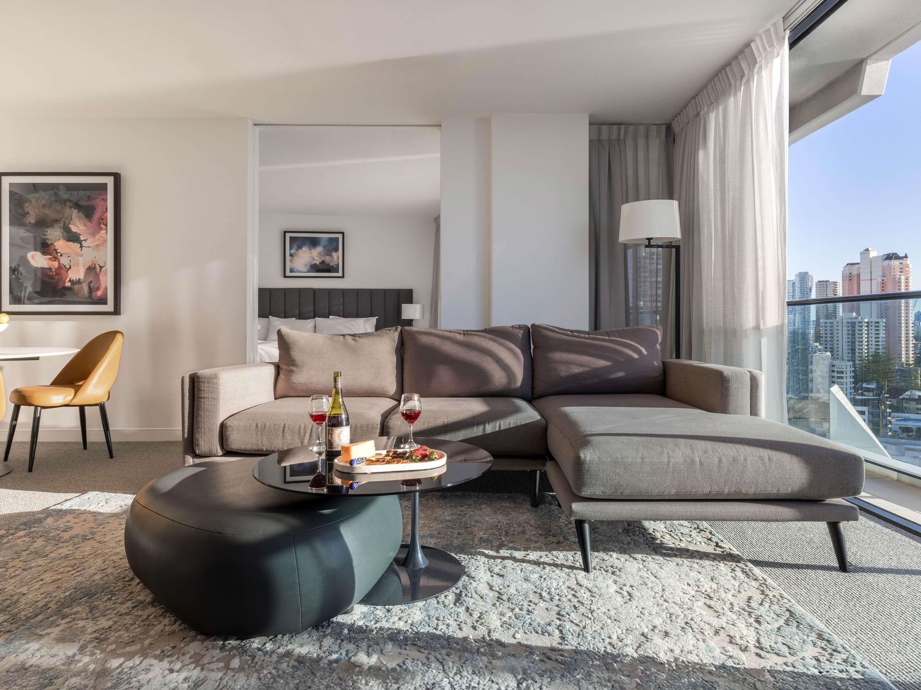 A modern hotel room with a sectional sofa, a round coffee table with a bottle and glasses, a chair, and a city view.