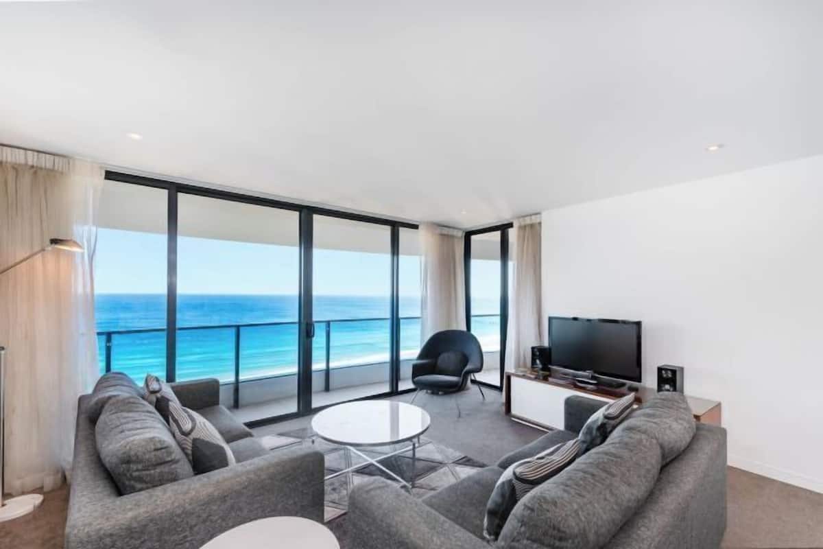 Una sala de estar moderna con un amplio ventanal con vistas al océano, un televisor de pantalla plana y una zona de estar con una mesita de centro.