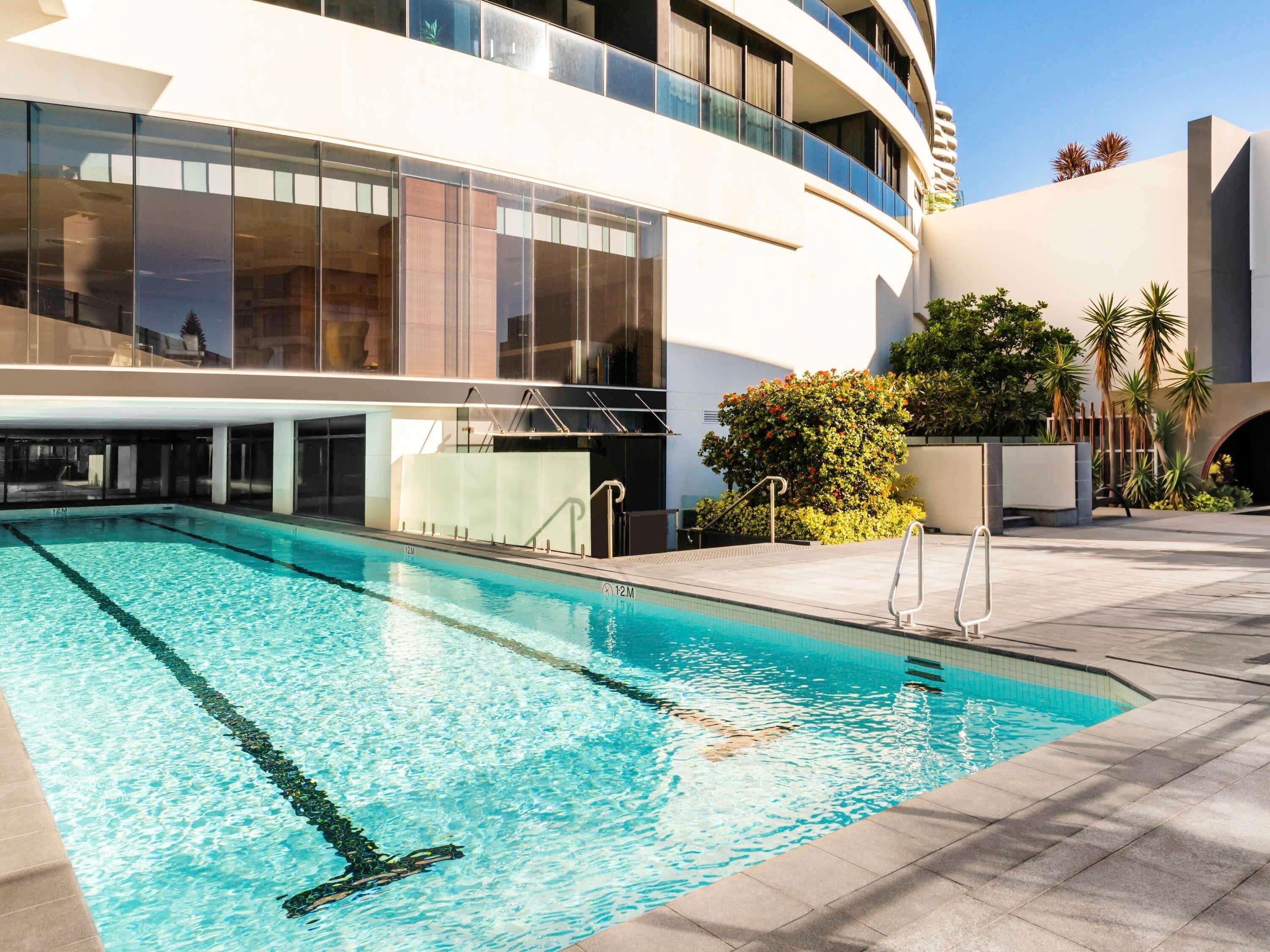Una piscina junto a un edificio moderno con amplios ventanales y un área ajardinada con palmeras.
