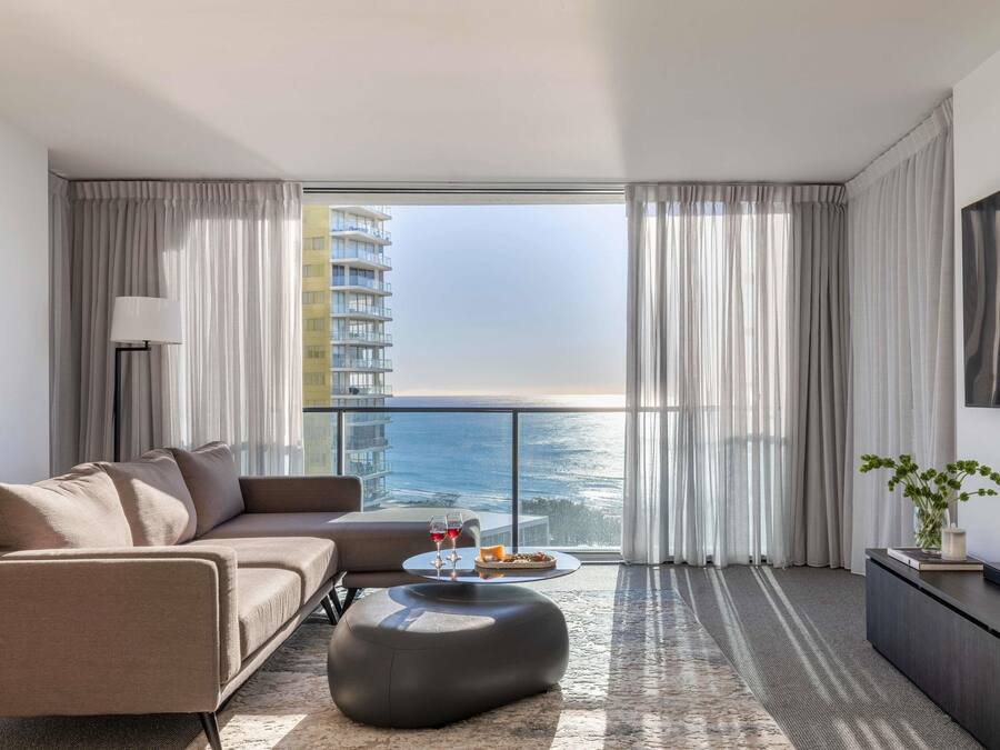 A modern living room with a sofa, ottoman, and coffee table, a floor lamp, and a view of the ocean through large windows.