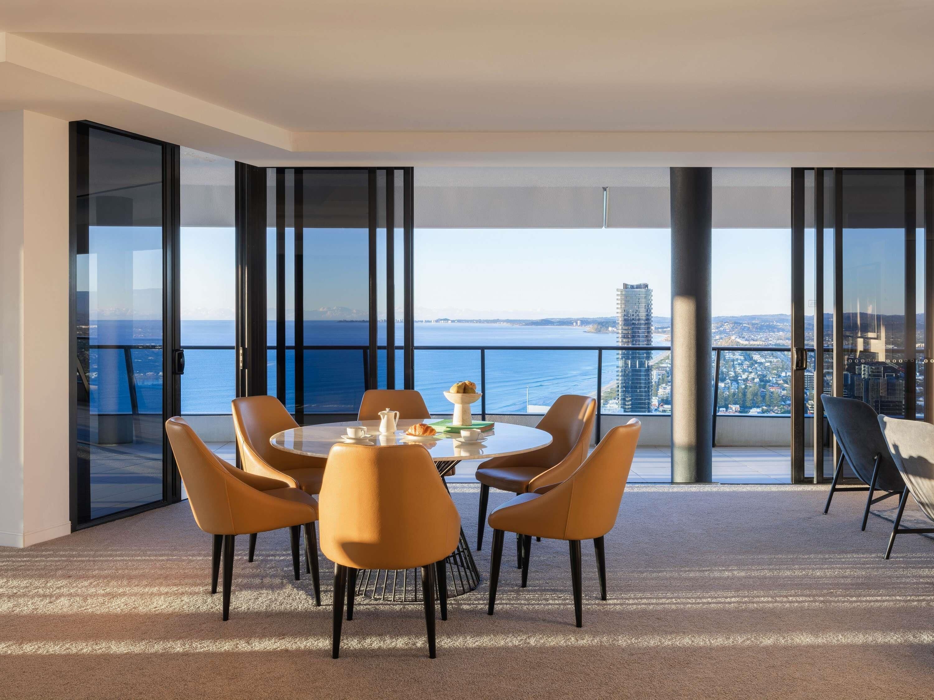 Un comedor moderno con una mesa dispuesta para cuatro, ventanas amplias que ofrecen vistas a la ciudad y al mar, y un balcón con vista al mar.