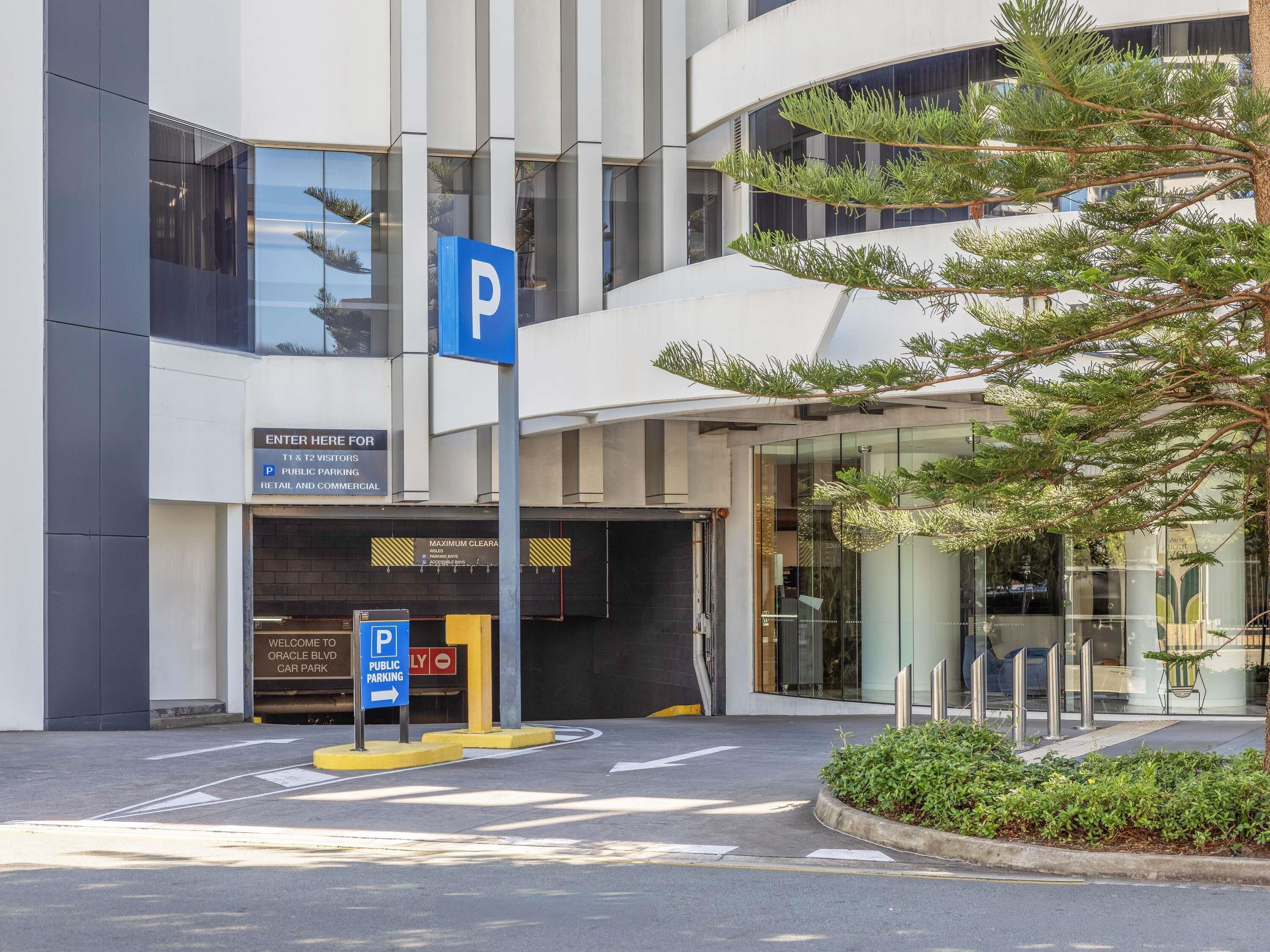 Entrada moderna de un edificio con un letrero de estacionamiento, un camino accesible y un área ajardinada con un árbol.