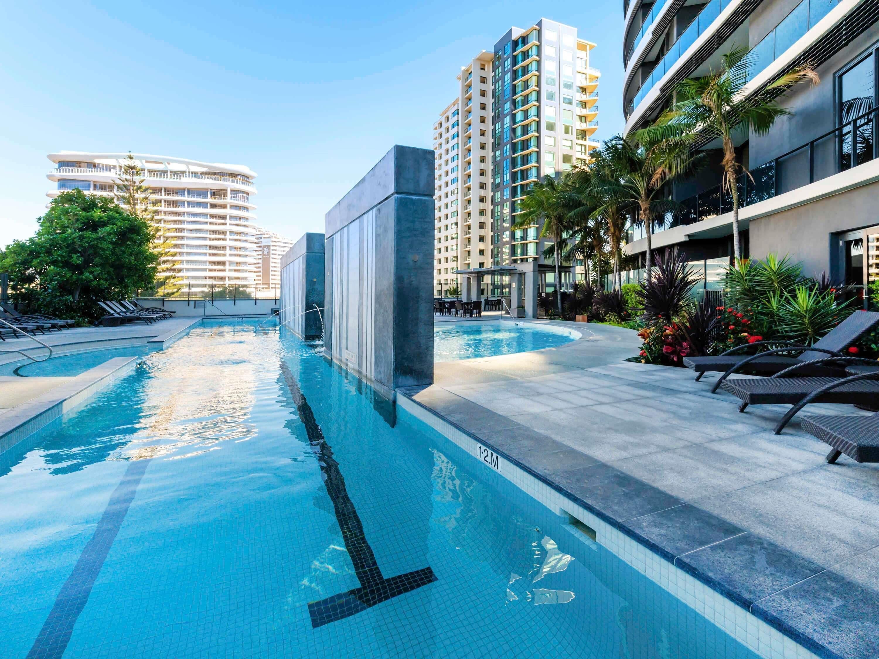 Una piscina con indicador de profundidad de 12 metros, rodeada de sillones de descanso y palmeras, con edificios altos al fondo.