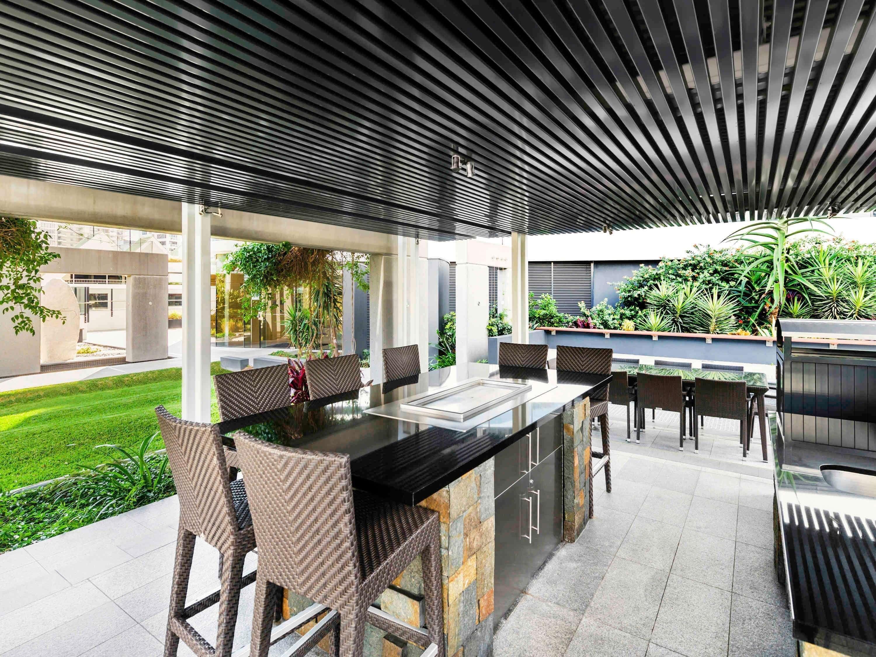 An outdoor dining area with a modern design, featuring a long table and chairs, surrounded by greenery and a covered seating area.
