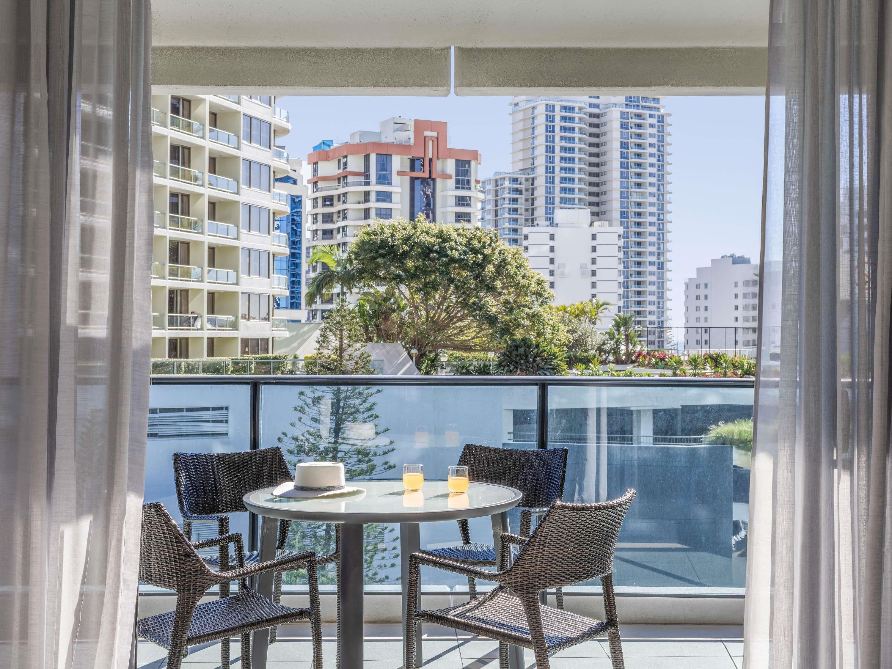 Un balcon avec des fauteuils en osier et une table, d’où l’on peut admirer un paysage urbain composé d’immeubles de grande hauteur et d’un arbre.