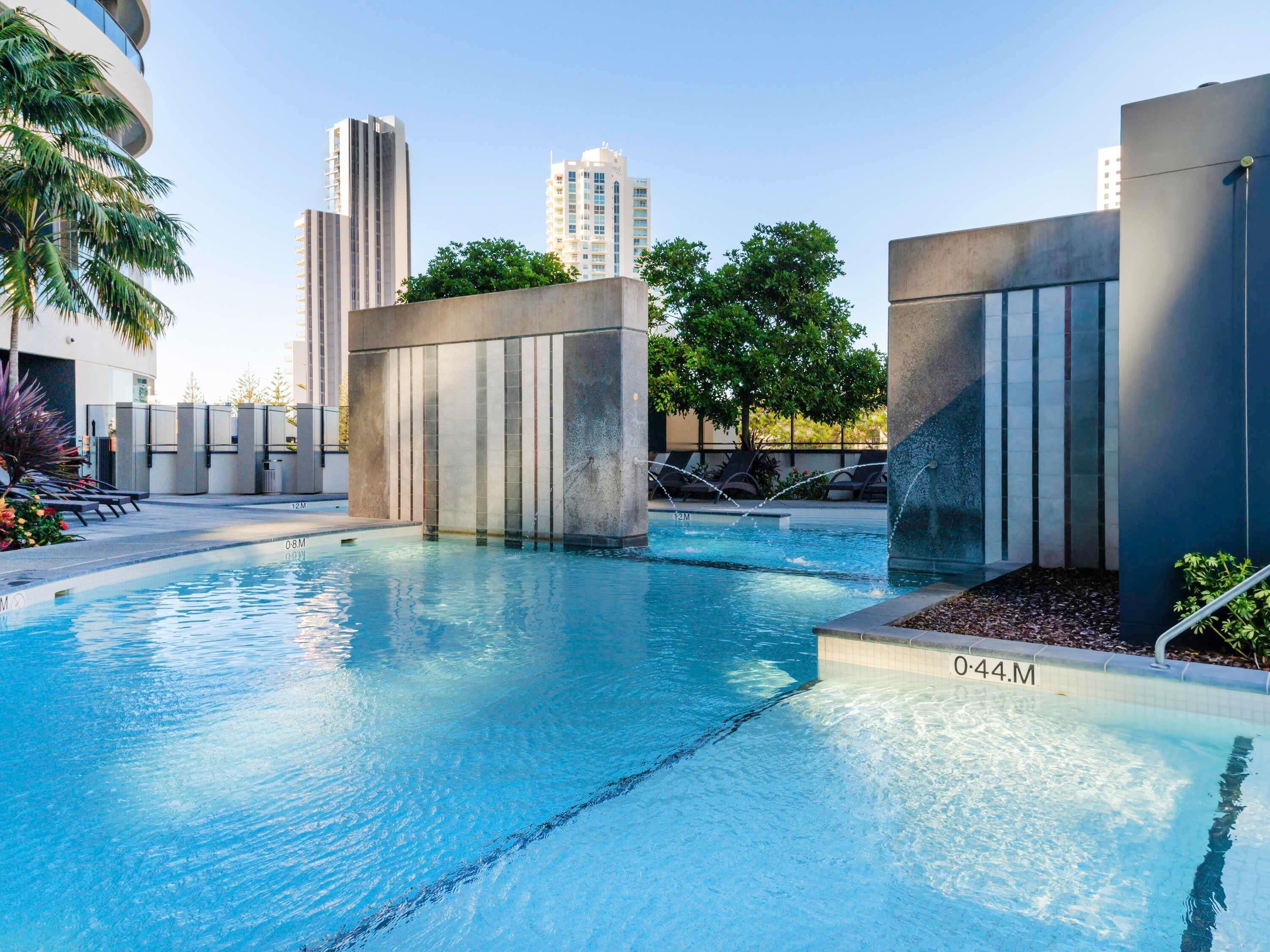 A modern pool area with clear water, surrounded by concrete structures and greenery, with tall buildings in the background.