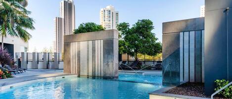 A modern pool area with clear water, surrounded by concrete structures and greenery, with tall buildings in the background.