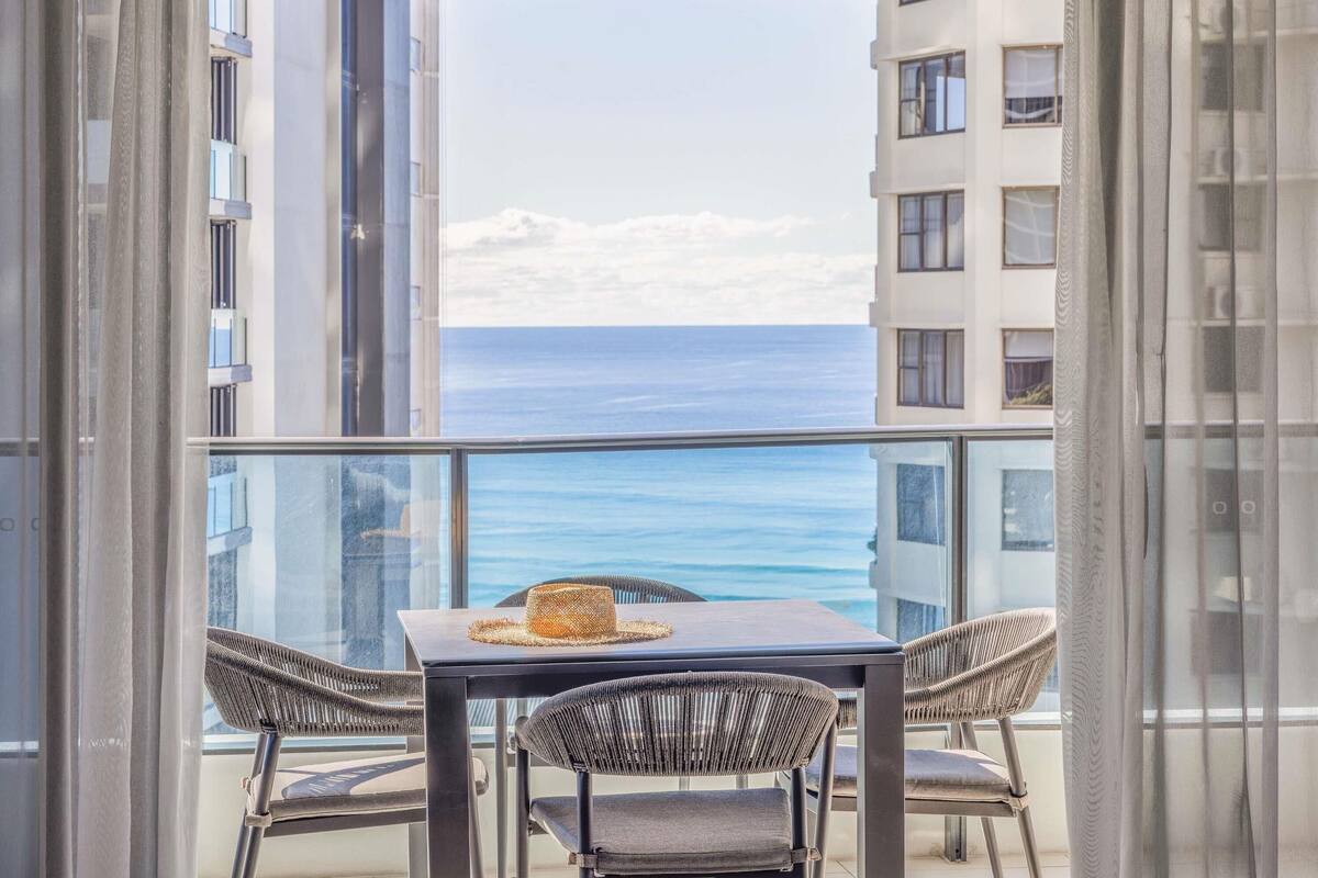 Un balcón con mesa y sillas, con vista a un cuerpo de agua y edificios.