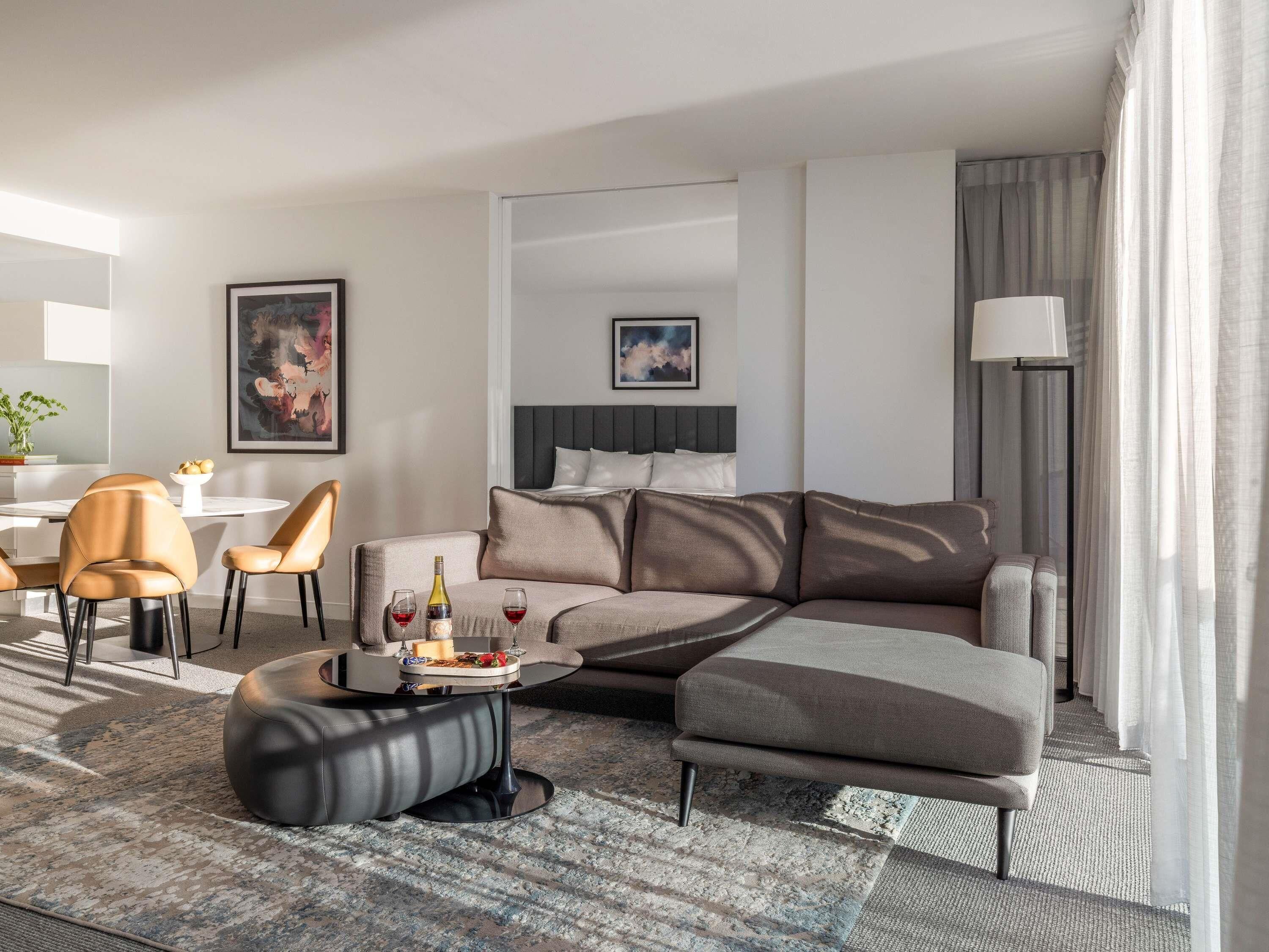 Ein modernes Hotelzimmer mit Ecksofa, rundem Couchtisch, Essbereich mit Stühlen, Bett mit Kopfteil und großem Fenster mit Vorhängen.
