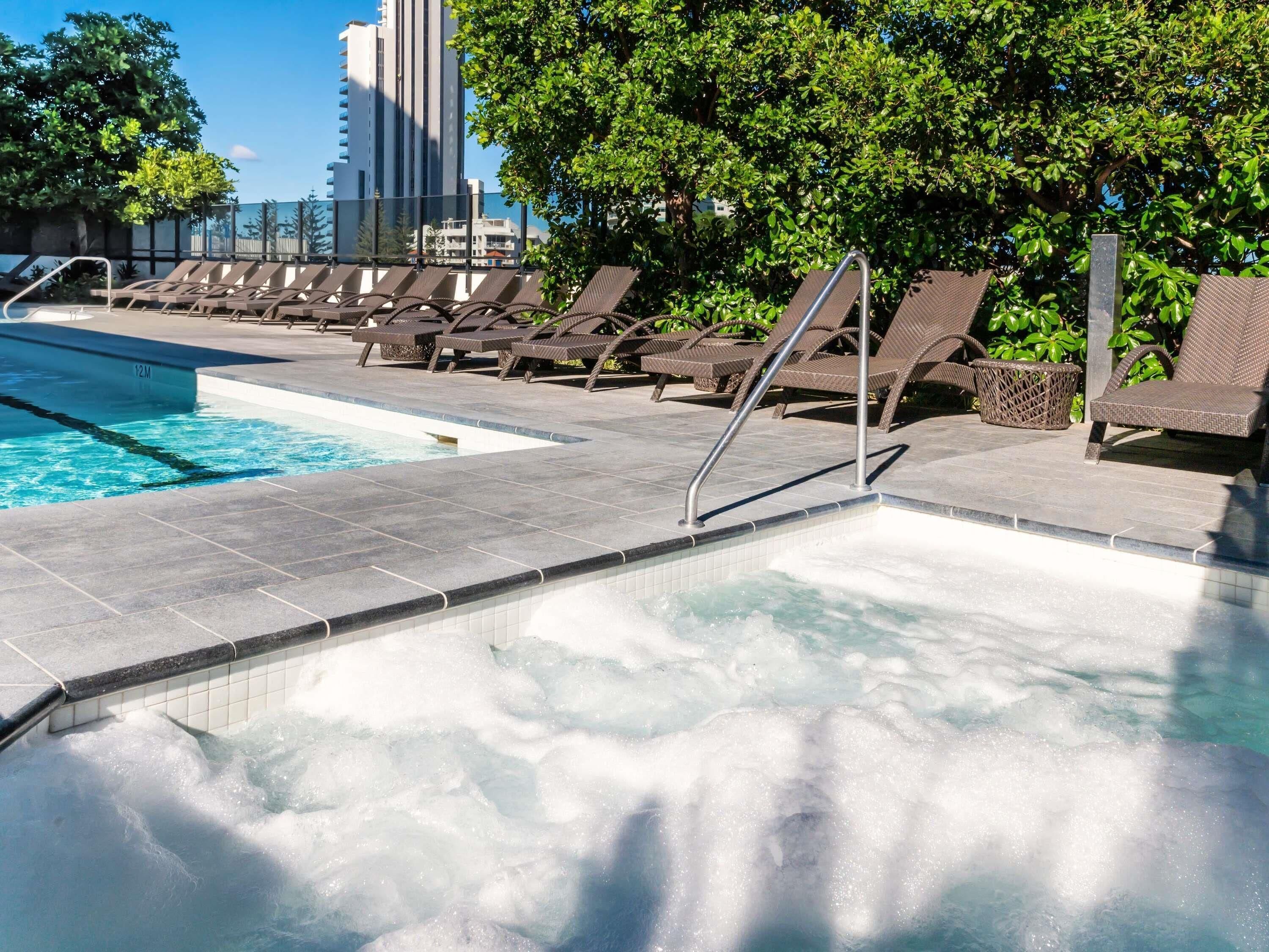 Una piscina con lettini, una vasca idromassaggio e uno sfondo urbano.