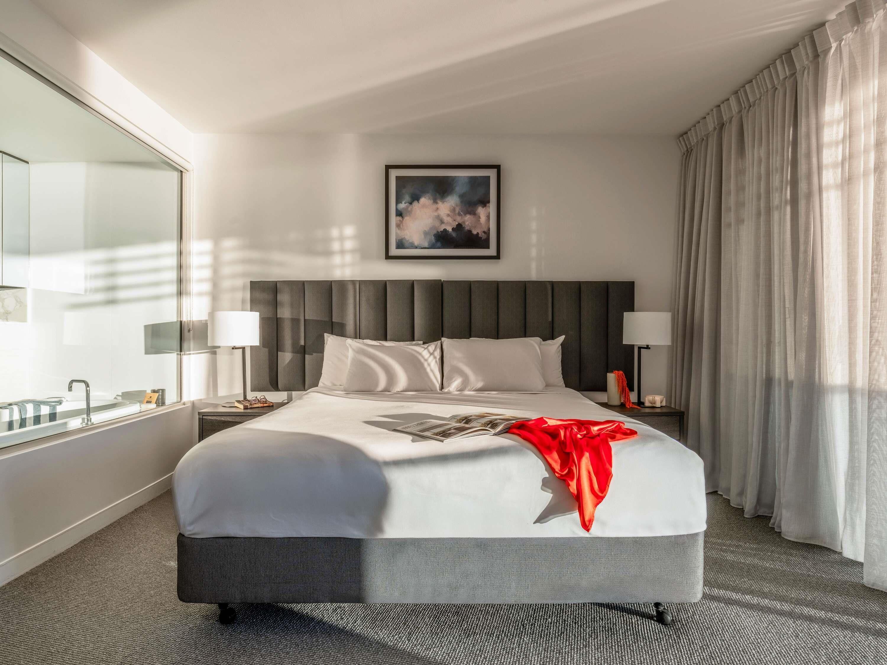 A hotel room with a large bed, a bedside table, a lamp, a window with curtains, and a framed picture on the wall.