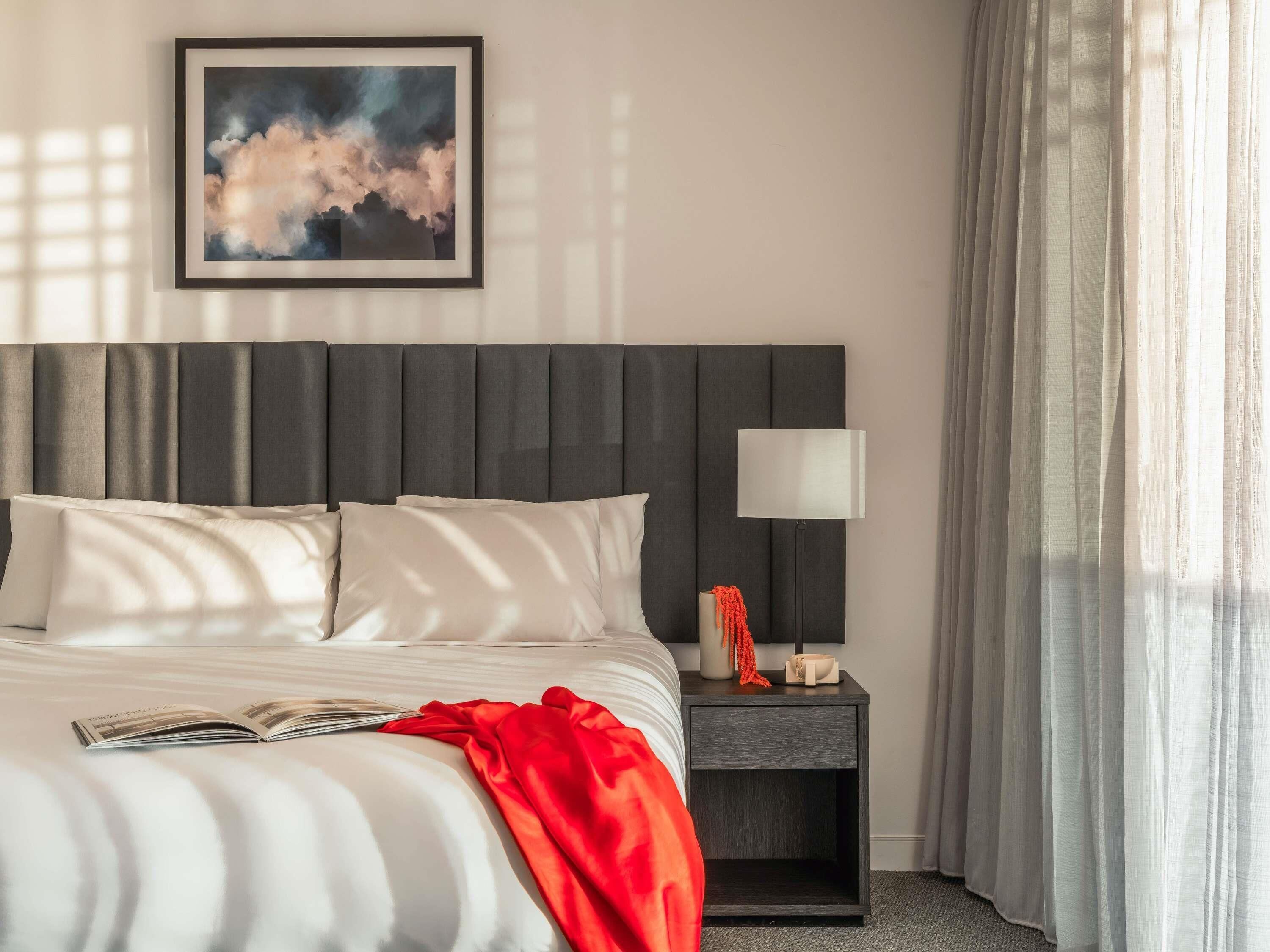 A neatly made bed with pillows, a bedside table with a lamp, a framed picture on the wall, and sheer curtains.