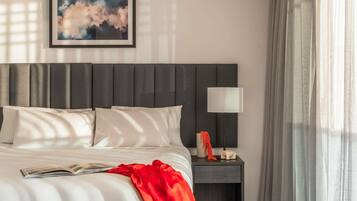 A neatly made bed with pillows, a bedside table with a lamp, a framed picture on the wall, and sheer curtains.