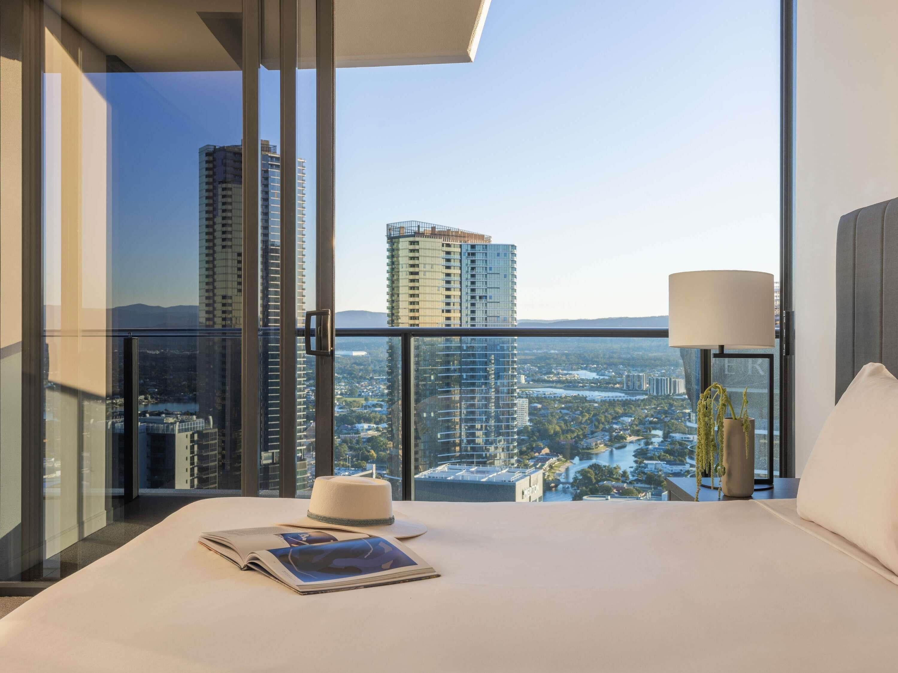 A hotel room with a large bed, a nightstand, a lamp, a hat, and a magazine. The room has a view of tall buildings and a cityscape.