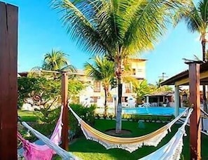 A hammock between wooden posts in a garden with a pool and palm trees. - Benko's Praia Hotel (Porto Seguro)