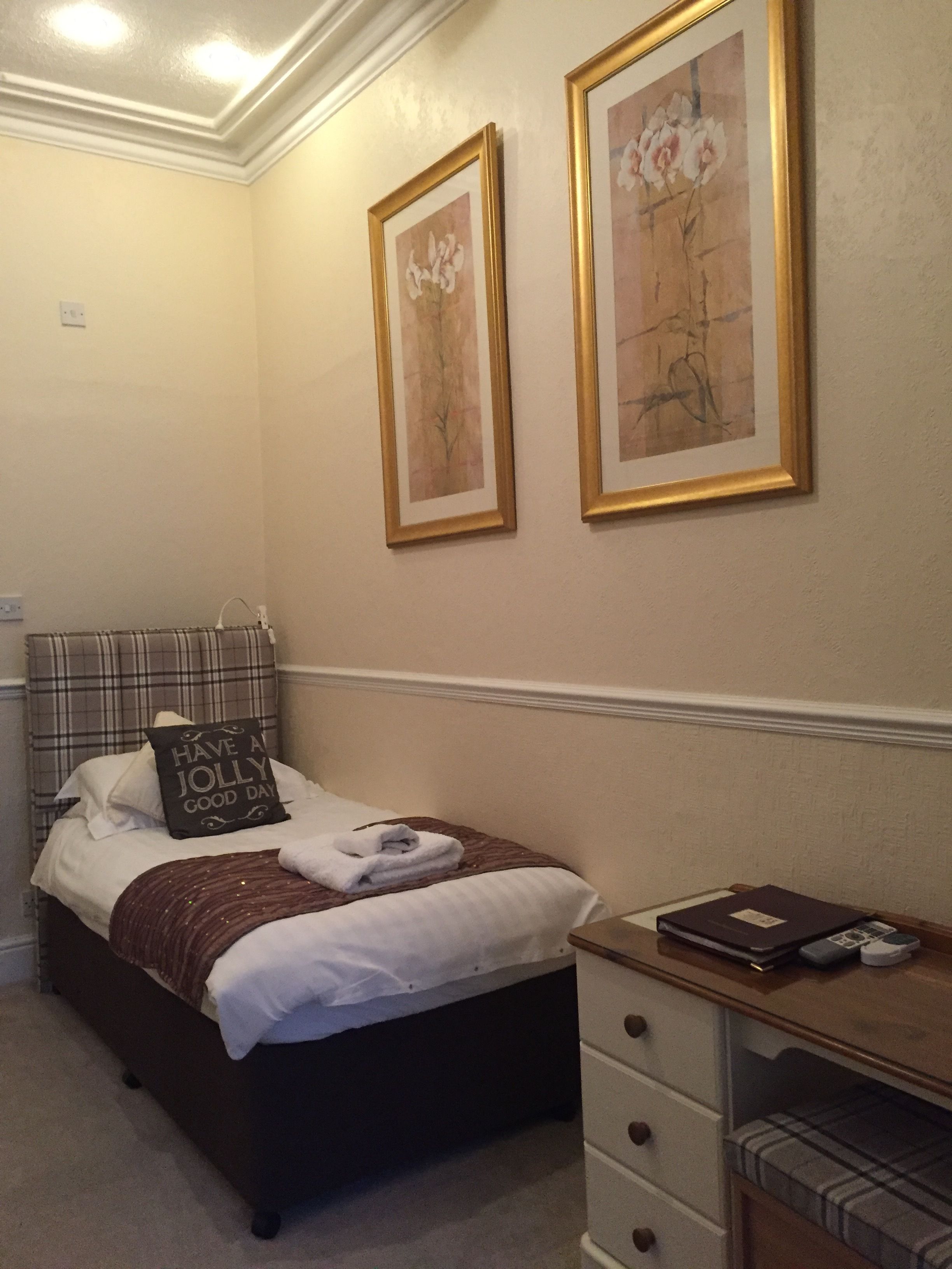 A neatly arranged bedroom with a bed, a bedside table with a book and a remote, and two framed floral paintings on the wall.