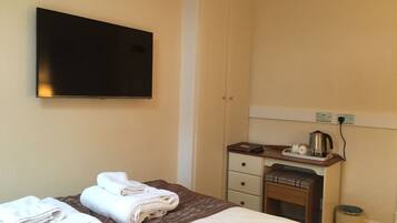 A hotel room with a bed, a TV mounted on the wall, a desk with a kettle, and a trash can.