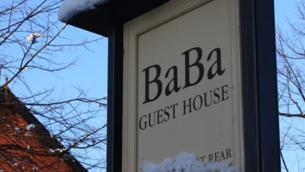 Naamplaatje voor 'BaBa Guest House', met een met sneeuw bedekte kap en kale bomen op de achtergrond.