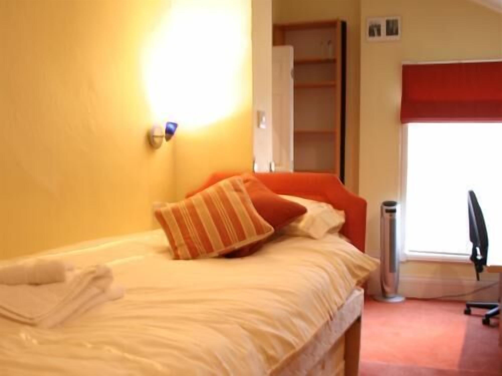 A bed with white bedding and orange pillows, a red window blind, and a trash can.