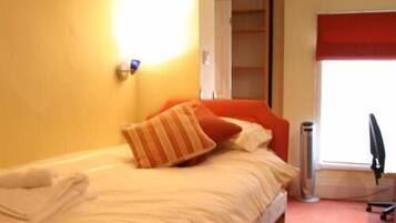 A bed with white bedding and orange pillows, a red window blind, and a trash can.
