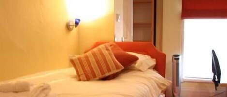 Een bed met witte lakens en oranje kussens, een rood raambedekking en een prullenbak.