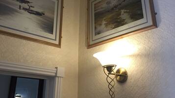 A room with two framed paintings on the wall, a lit wall sconce, and a mirror reflecting a door with a framed picture.