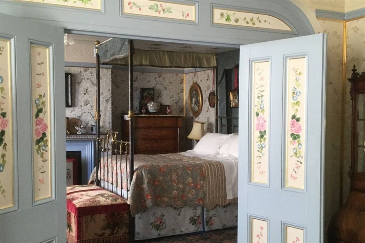 A bedroom with a four-poster bed, a wooden dresser, and floral wallpaper.