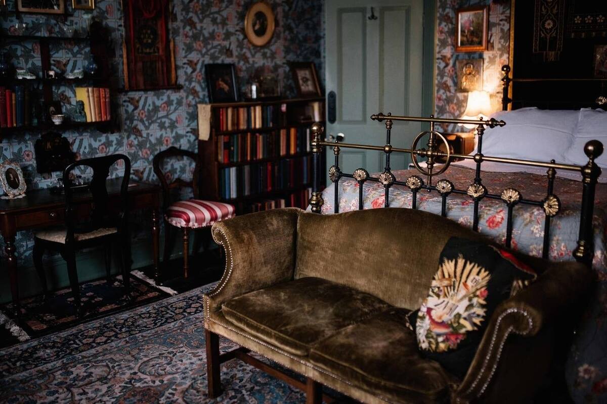 A vintage-style room with a patterned carpet, a wooden desk, a striped chair, a bookshelf, and a bed with a floral headboard.