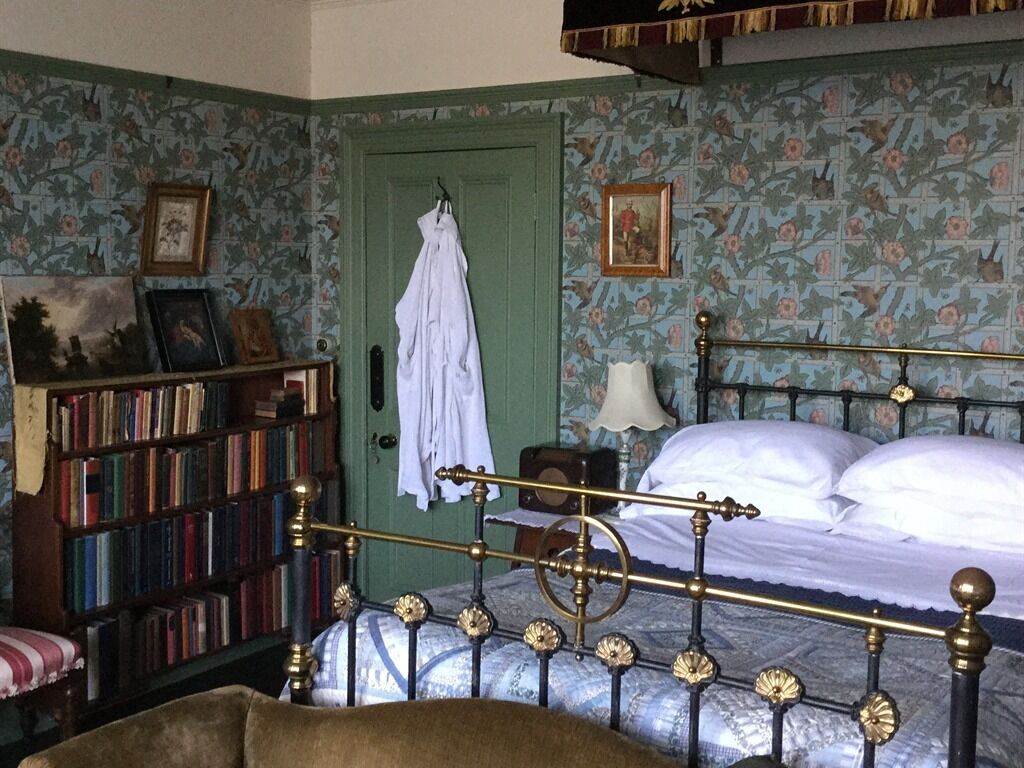 Een slaapkamer met een hemelbed, een boekenkast, een deur met een jas er aan, en bloemenbehang.